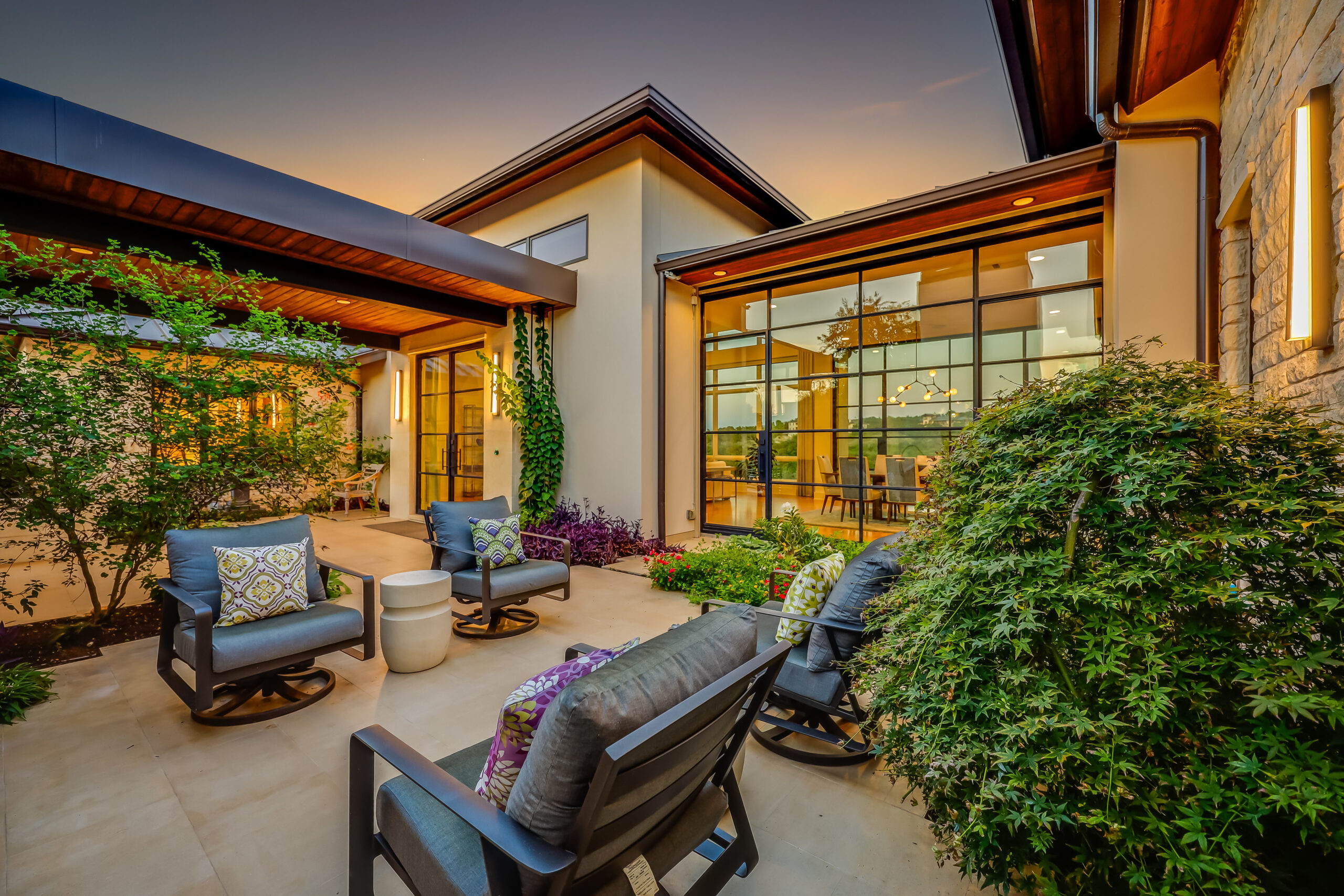 Exterior view of a modern luxury home at dusk, featuring large glass windows and doors, with an inviting outdoor seating area including two chairs with cushions and a small table. The architecture showcases clean lines, a mix of materials with warm lighting that enhances the home’s design. Lush greenery and well-maintained landscaping add to the property’s curb appeal, suggesting a high-end real estate offering in Austin or Colorado.