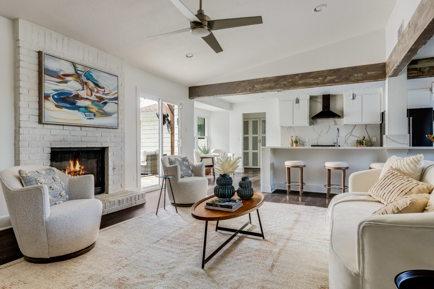 A modern living room with a white brick fireplace, two beige armchairs, a beige sofa with patterned and solid cushions, a wooden coffee table with decorative vases, and an open kitchen with a breakfast bar